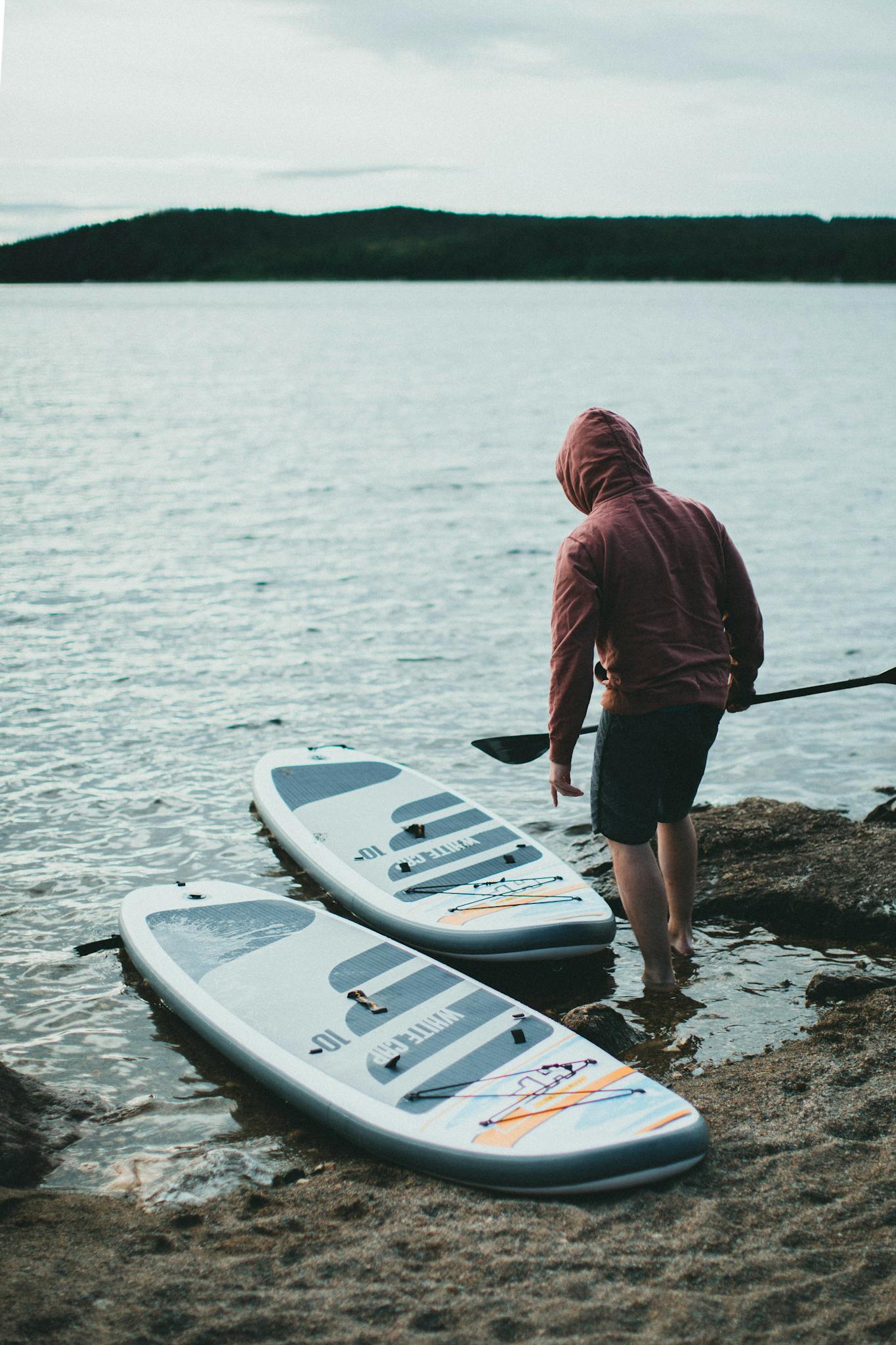 A person in a hoodie prepares paddle boards by a serene lake shore under cloudy skies.